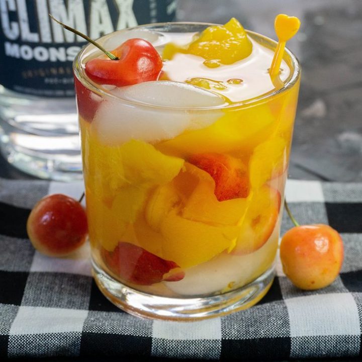 Cocktail with rainier cherries on black plaid napkin with moonshine bottle in the back