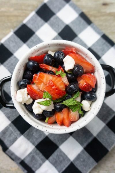 July 4th Fruit Salad with strawberries, blueberries, and mozzarella pearl cheese in a whote bowl with black speckles on a black and white plaid napkin